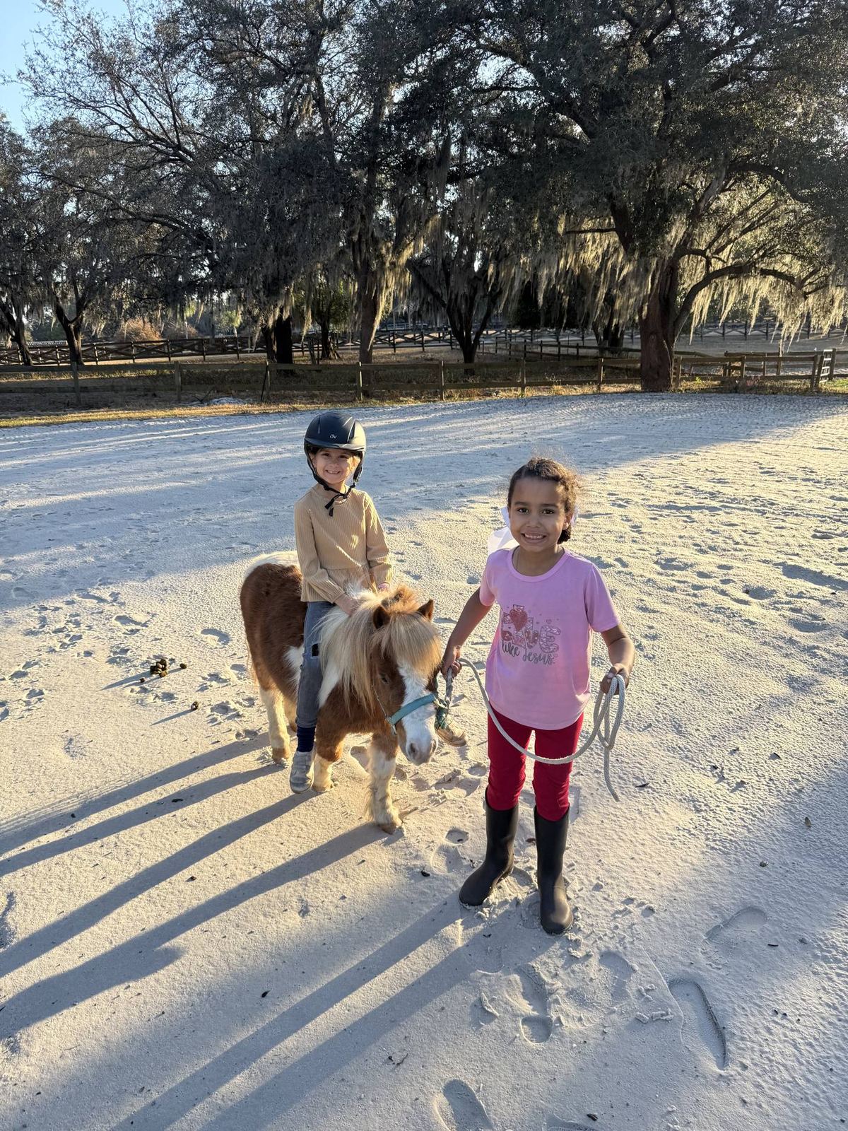 Two children in a sandy arena, one riding a small pony while the other leads it